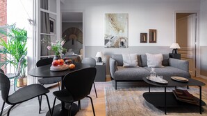 A modern living room with a grey sofa, black chairs, a dining table with fruit, and a large abstract painting on the wall. - CASA CALABRIA 2 BEDROOM 4 PAX (Barcelona)
