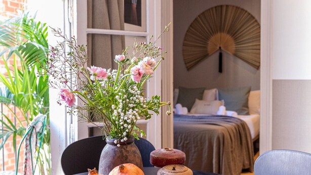A dining table with a vase of flowers, plates, and a pomegranate. A bed and a fan are visible in the background. - CASA CALABRIA 2 BEDROOM 4 PAX (Barcelona)