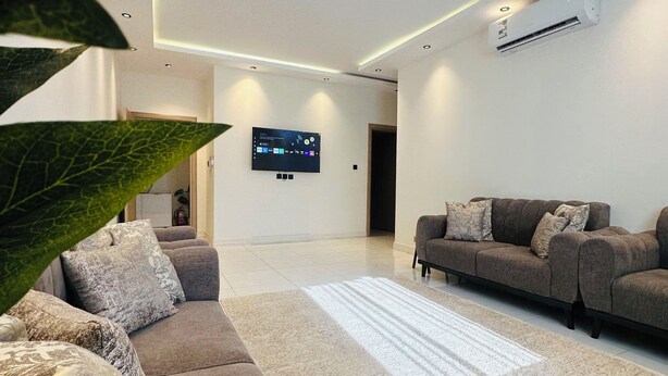 A modern living room with a grey sofa set, a flat-screen TV mounted on the wall, and an air conditioning unit. - Spacious & New 4-bedroom apartment in peaceful Madinah with AC, Fast-WiFi (Madinah)
