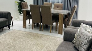 A modern living room with a grey sofa, a dining table with six chairs, a potted plant, and a large window with curtains. - Spacious & New 4-bedroom apartment in peaceful Madinah with AC, Fast-WiFi (Madinah)