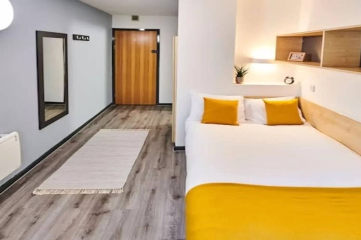 Une chambre d’hôtel avec un plancher en bois, un lit avec des oreillers jaunes, une table de chevet avec une lampe et une petite étagère avec une plante.