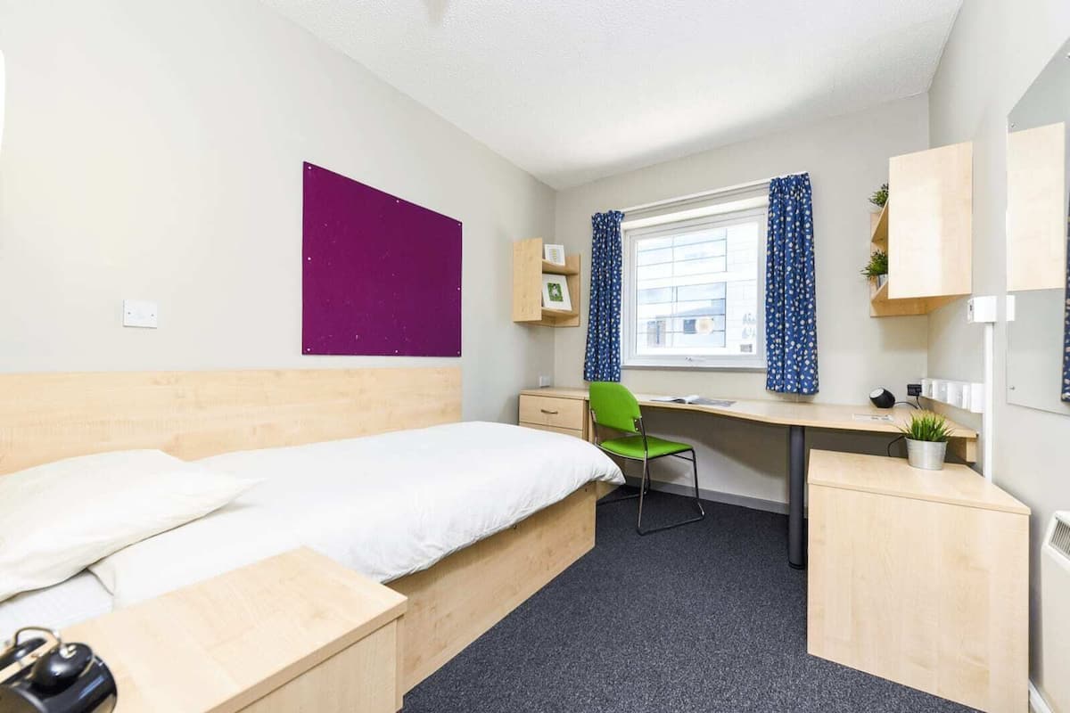 Une chambre d’hôtel de taille réduite, comprenant un lit, un bureau, une chaise et une petite kitchenette.