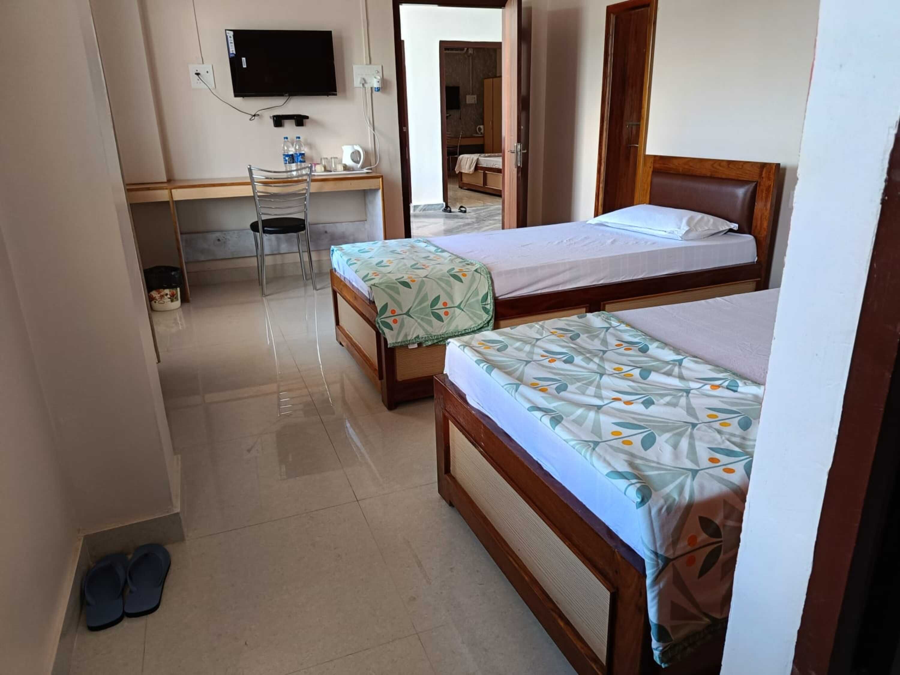 Et hotelværelse med to senge, et skrivebord, en stol, et fjernsyn og et par badesko på gulvet.