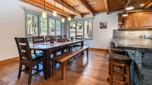 Una mesa de comedor de madera con bancos y taburetes de bar, una cocina con armarios de madera y ventanas grandes con vistas a árboles.