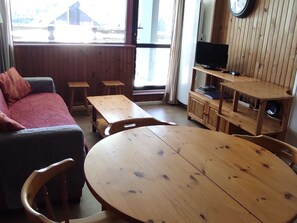 A room with a round wooden table, a sofa, two wooden benches, a television on a wooden stand, and a window with a view of buildings.