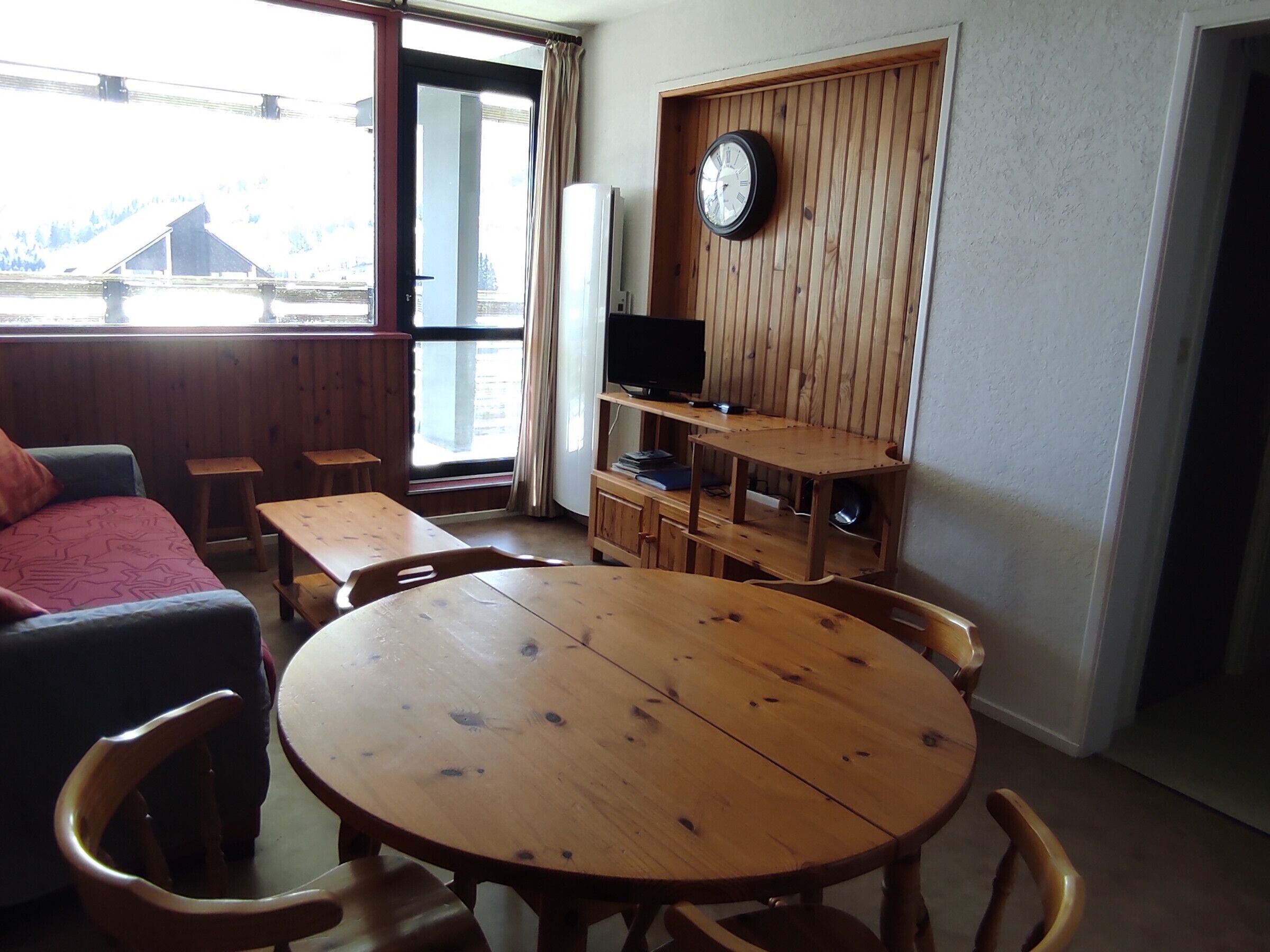 A room with a round wooden table, chairs, a sofa, a TV on a wooden shelf, and a clock on the wall.