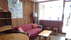 A room with a wooden floor, a red patterned sofa, a wooden table, two wooden stools, a lamp, and a painting on the wall.