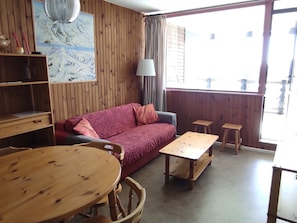 A room with a wooden floor, a red patterned sofa, a wooden table, two wooden stools, a lamp, and a painting on the wall.