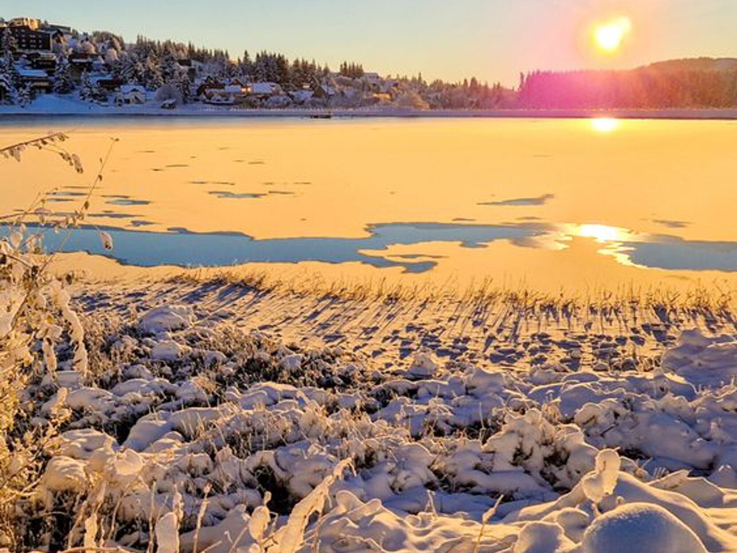 Winterliche Landschaft mit einem zugefrorenen See, verschneitem Boden und einem Gebäude im Hintergrund.