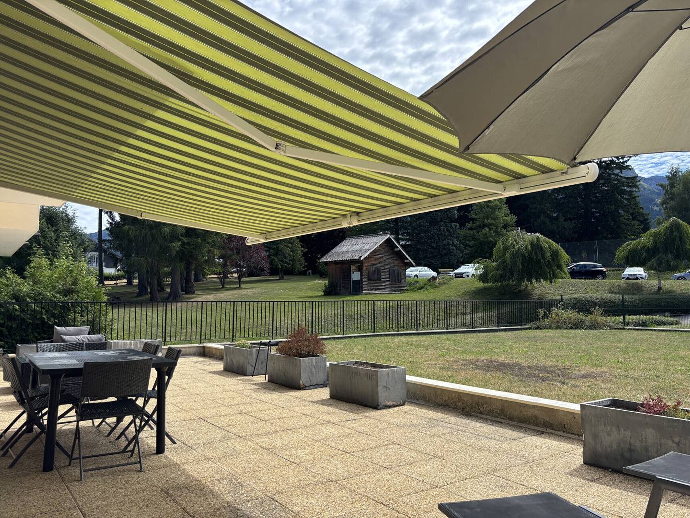 An outdoor patio area with a table and chairs, a covered seating area with a striped awning, and a view of a grassy area with trees and a small building.