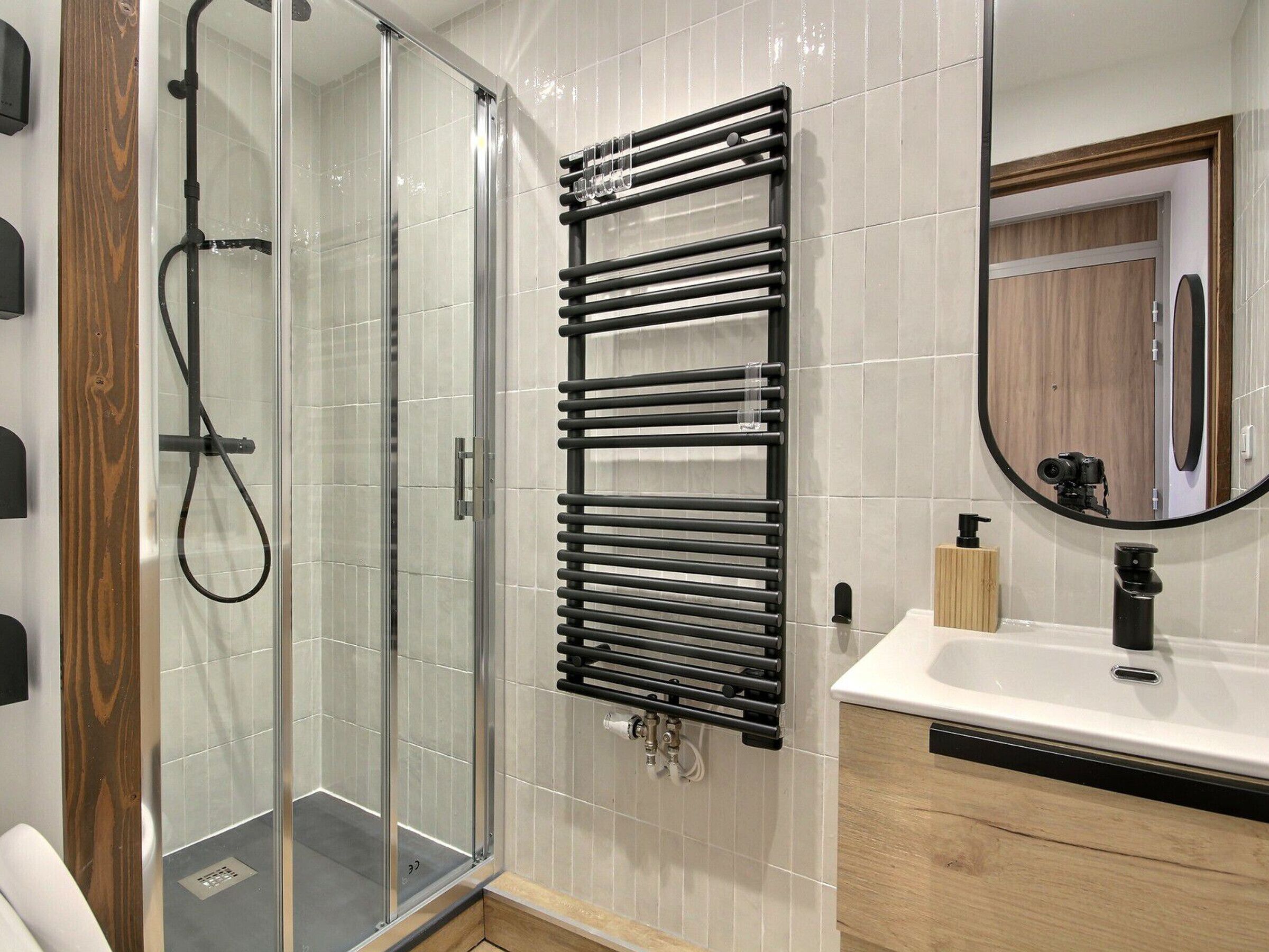 Un baño moderno con cabina de ducha de vidrio, toallero negro, lavamanos blanco con grifo negro y tocador de madera.