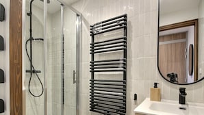 Un baño moderno con cabina de ducha de vidrio, toallero negro, lavamanos blanco con grifo negro y tocador de madera.