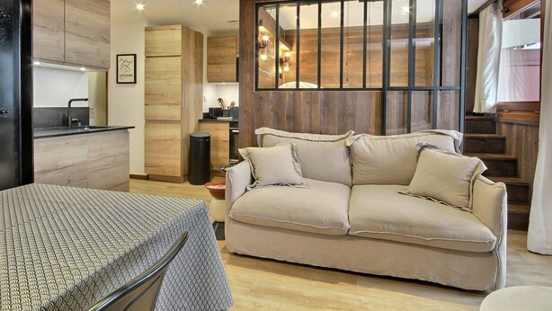 Una sala de estar moderna con un sofá color beige, una mesa de comedor con mantel estampado y una cocina con armarios de madera y un cesto de basura negro.