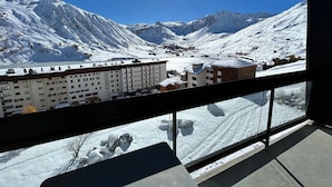 Paisagem nevada de montanhas com céu claro e o sol brilhando. Há edifícios com telhados cobertos de neve e uma varanda com vista para as montanhas.