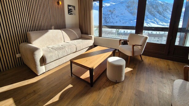 Uma sala de estar moderna com um sofá, uma mesa de centro e uma poltrona, com grandes janelas e vista para as montanhas.
