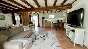 A spacious living area with a sofa, coffee table, and TV. It features exposed wooden beams, a kitchen with a dining area, and a large window.