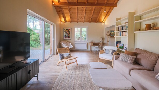 A spacious living room with a large TV, comfortable seating, a wooden ceiling, and large windows offering a view of greenery. - Villa Madonne *Pool *Beach *7 sleeps ☀️ (Lège-Cap-Ferret)