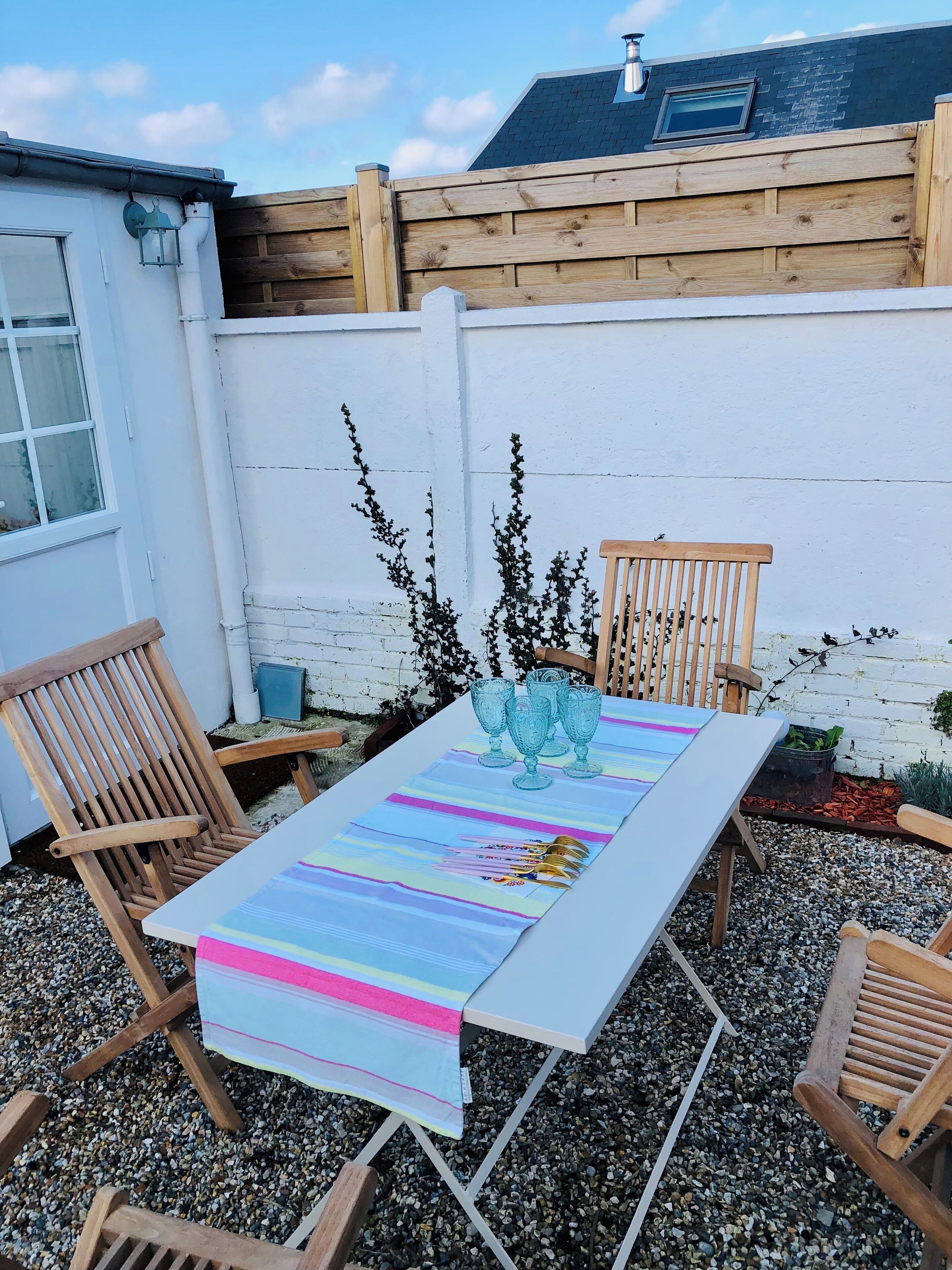 Une terrasse avec une table dressée pour quatre personnes, entourée de chaises en bois, d’un mur blanc et d’une clôture en bois.