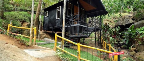 A black cabin with a wooden deck and stairs, surrounded by a yellow safety fence and lush greenery.