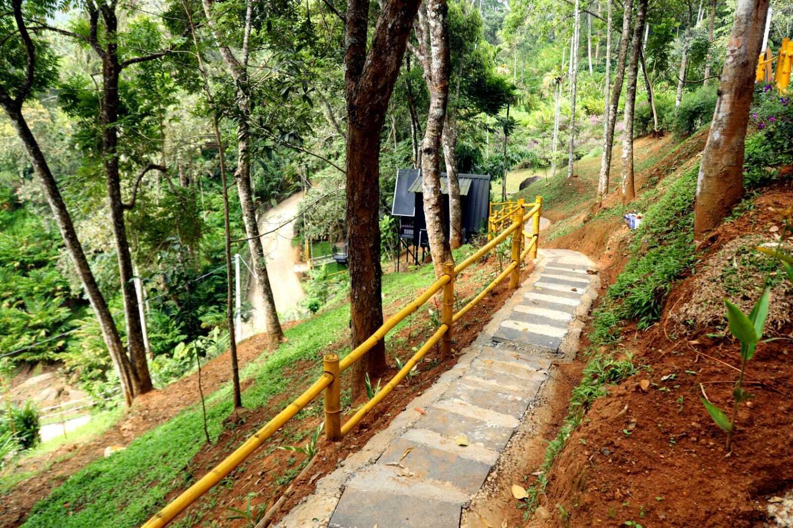 Uma escadaria de pedra com corrimão amarelo serpenteia por um declive, cercada por vegetação densa e árvores.