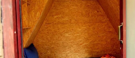 Pequeña habitación triangular en el ático con una cama individual, un colchón azul y una manta doblada.