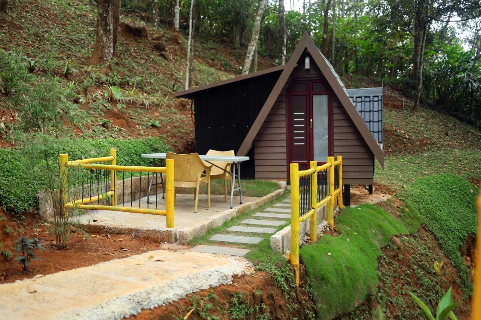 A small wooden cabin with a red door, surrounded by a yellow fence and a concrete pathway leading to a table and chairs.