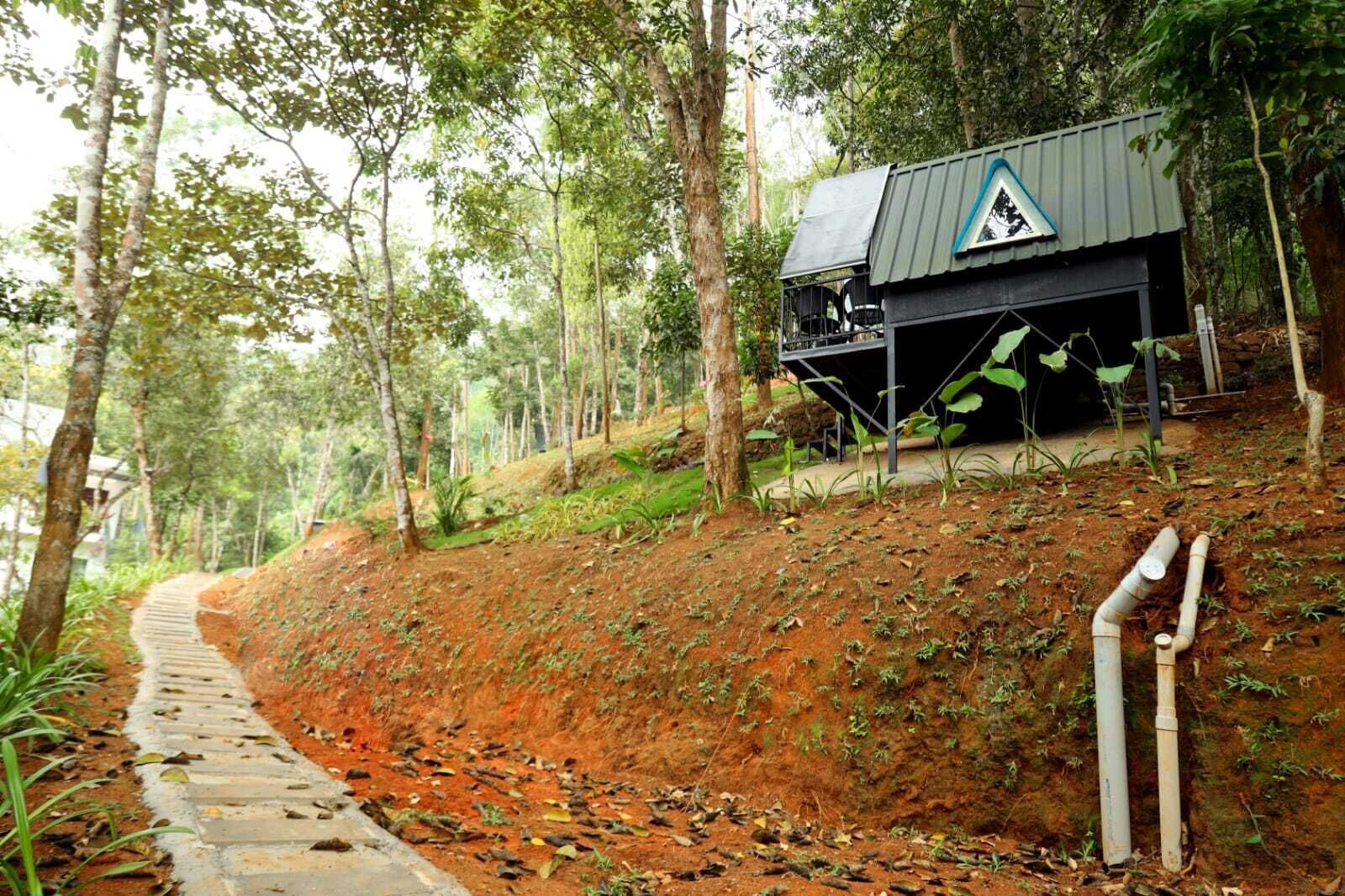 Uma cabana rústica com telhado de metal e uma janela triangular, cercada por uma área arborizada com uma trilha de terra que a leva.
