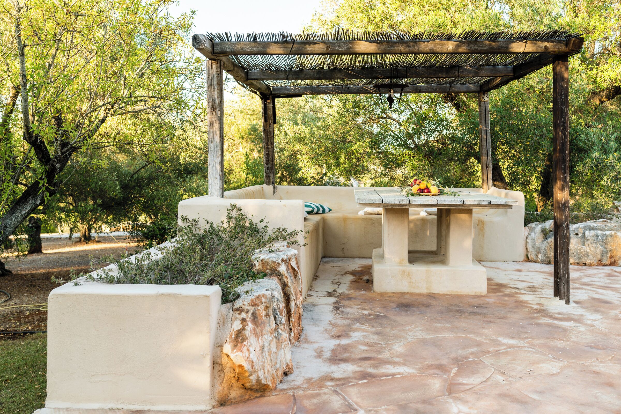 An outdoor patio with a pergola, stone seating, and a table with fruits.