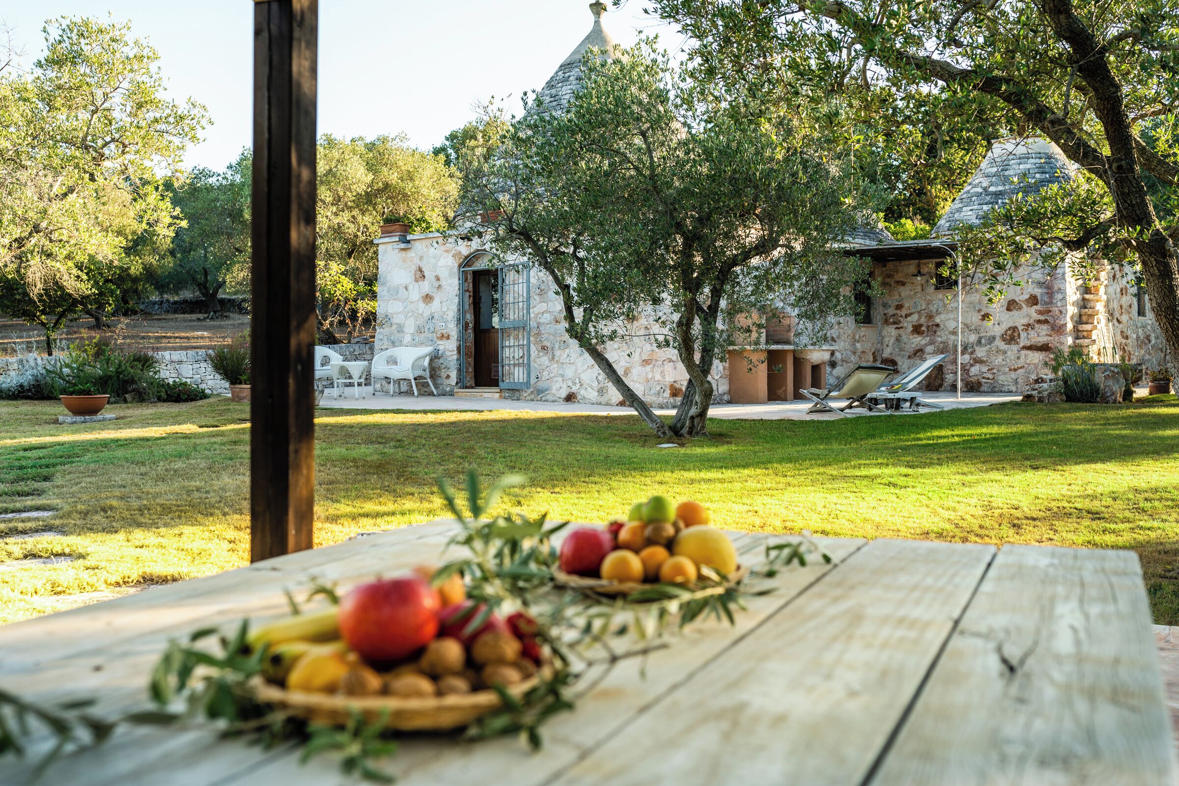 A rustic stone building with a tiled roof, a wooden table with fruit and greenery, and a grassy yard with a tree and a chair.
