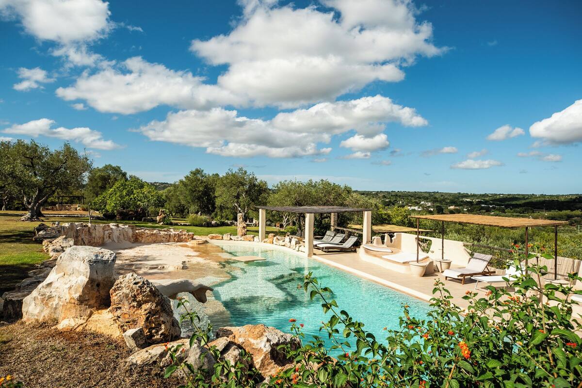 A pool area with lounge chairs and a pergola, surrounded by greenery and a clear sky.