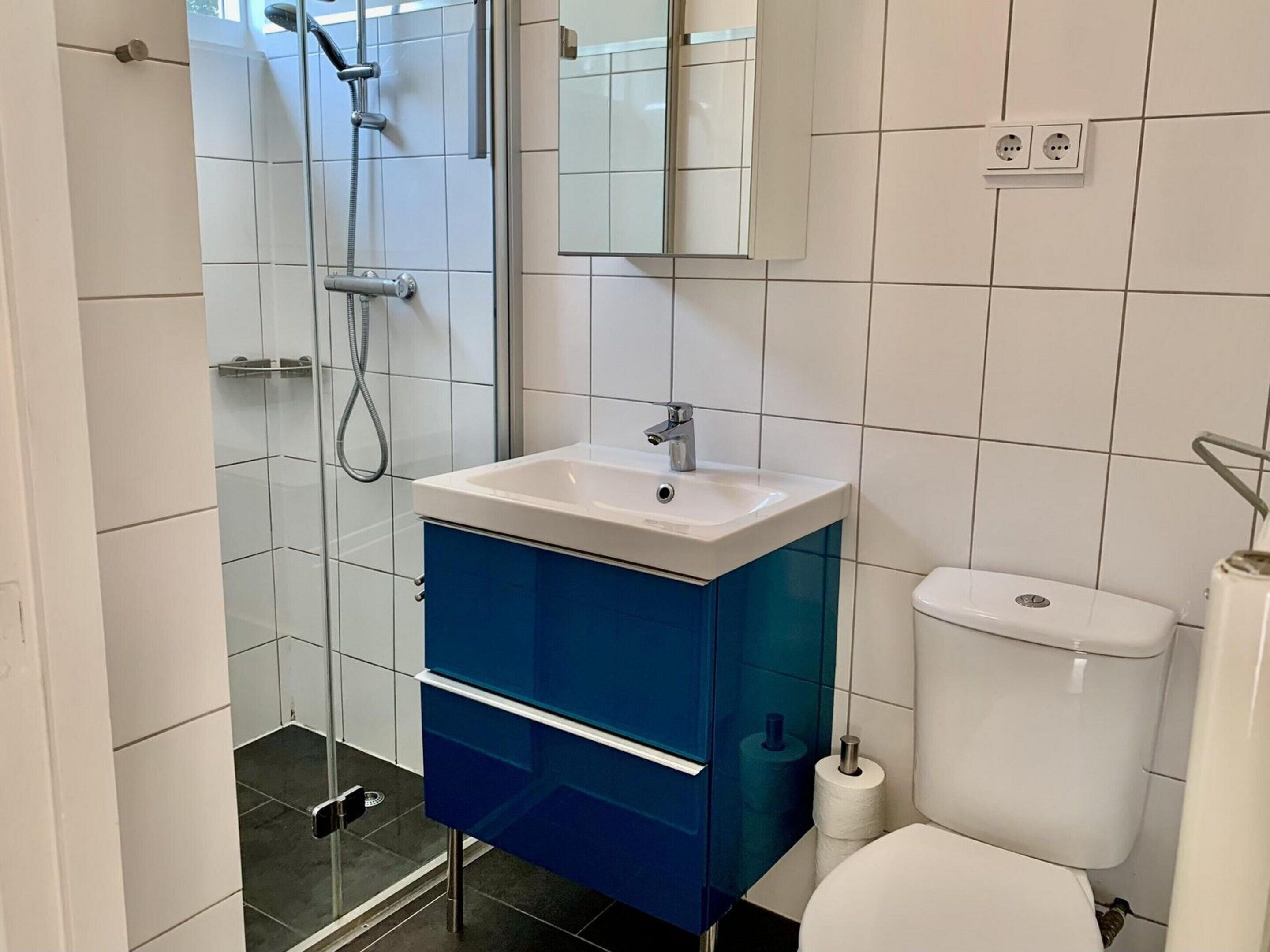 A bathroom with a glass shower enclosure, a white sink with a blue cabinet, and a white toilet.