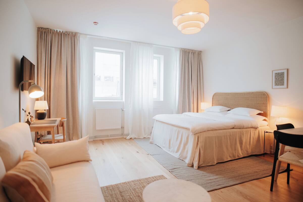 A well-lit bedroom with a large bed, a bedside table, a chair, and a window with curtains.