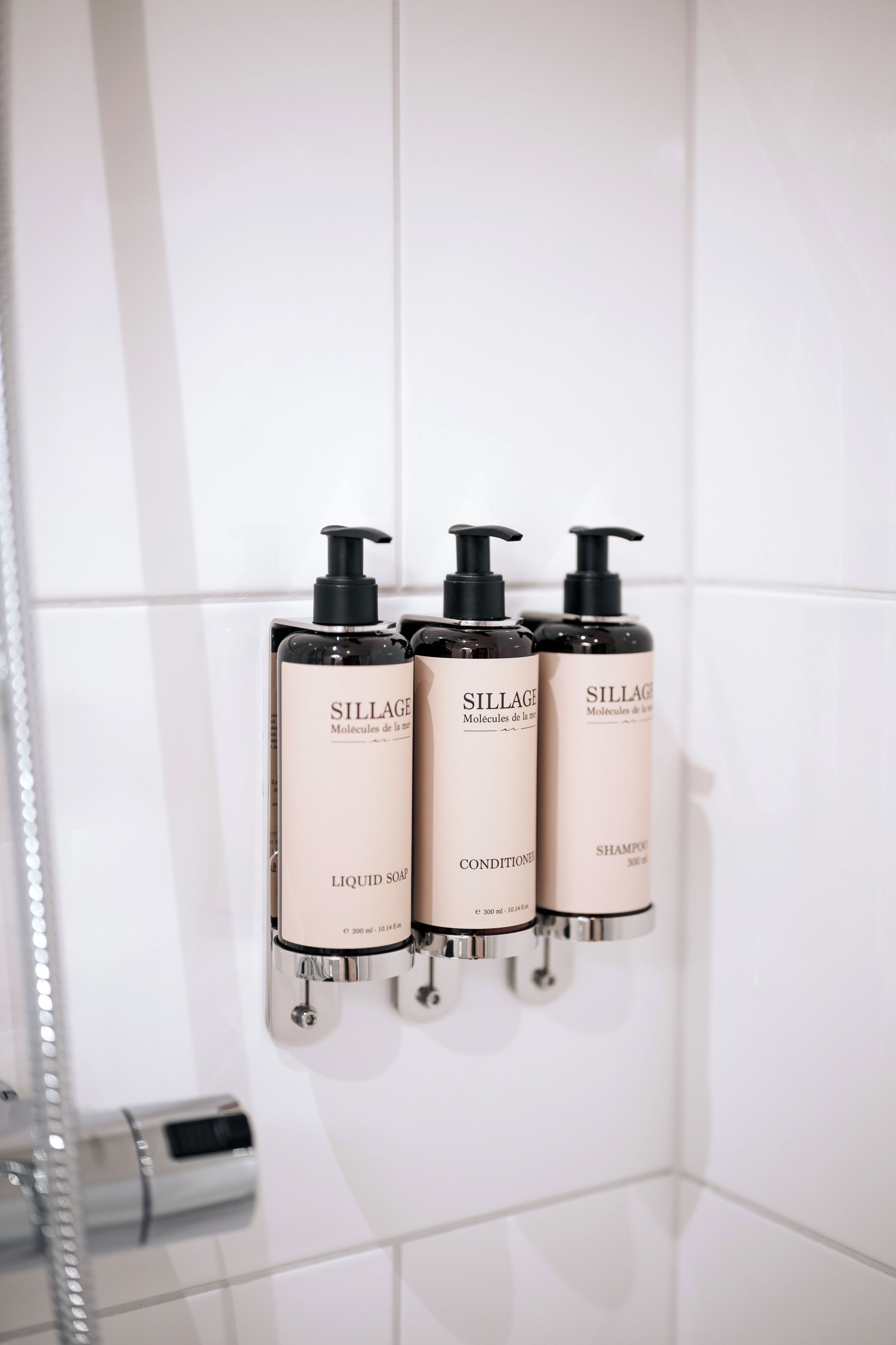 Tres botellas de champú Sillage montadas sobre una pared de azulejos blancos en un baño.