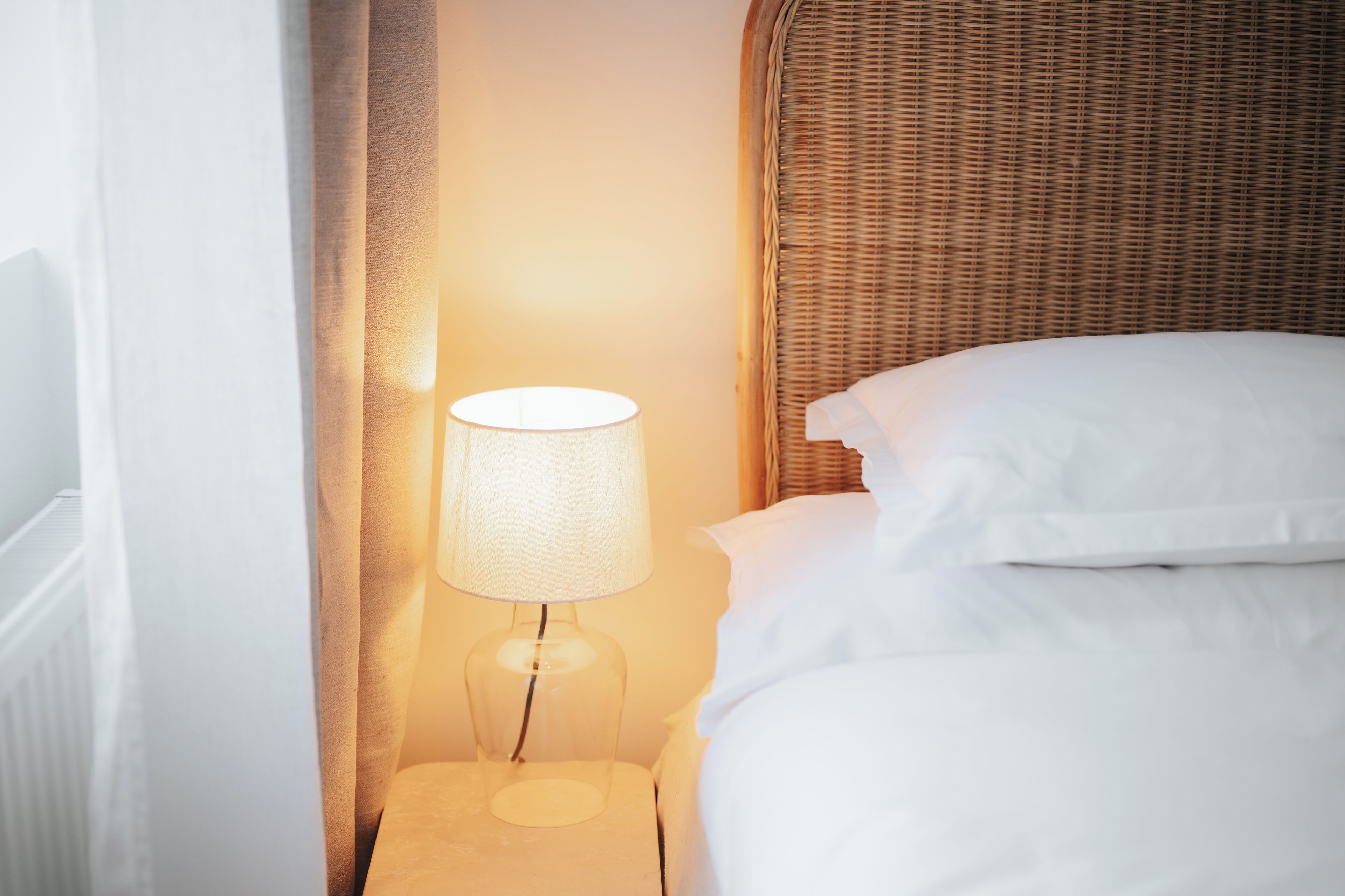 Ein ordentlich gemachtes Bett mit weißen Leinen, einer Glas Tischlampe und einem Rattan-Kopfstück.