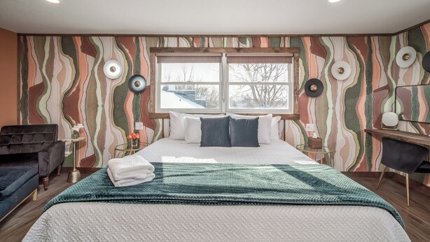 A modern bedroom with a large bed, a view of the outdoors through a window, and a unique wavy-patterned wall. - Extended Stay | Spacious Kitchen Studio | Free Parking (Prescott)