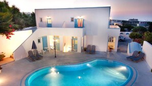 A modern building with a swimming pool, lounge chairs, and a view of the surrounding area. - Villa Pelekanos Santo (Santorini)