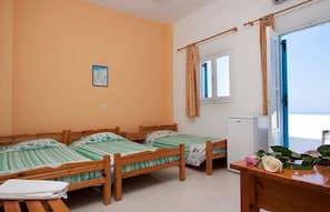 A room with three wooden beds, a small table with a vase of flowers, and a view of the sea through an open door.