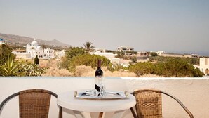 A table with a bottle and glass on a terrace overlooking a coastal town with white buildings and a mountain in the background.