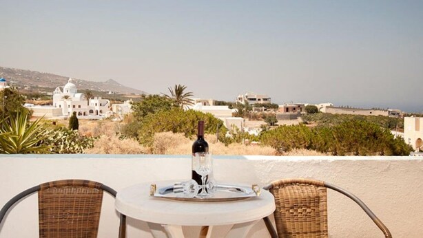 A table with a bottle and glass on a terrace overlooking a coastal town with white buildings and a mountain in the background. - Villa Pelekanos Santo (Santorini)