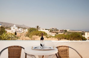 A table with a bottle and glass on a terrace overlooking a coastal town with white buildings and a mountain in the background. - Villa Pelekanos Santo (Santorini)