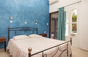 A bedroom with a bed, a nightstand, a window with curtains, and an air conditioning unit. - Villa Pelekanos Santo (Santorini)