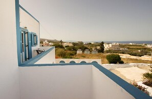 Un edificio blanco con detalles y contraventanas azules, con vista a un paisaje costero con casas y vegetación.
