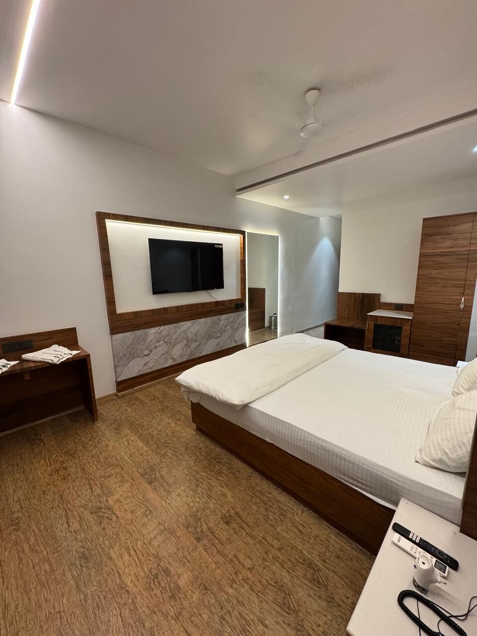 A modern hotel room with a large bed, a flat-screen TV mounted on the wall, a wooden nightstand, and a desk with a lamp.