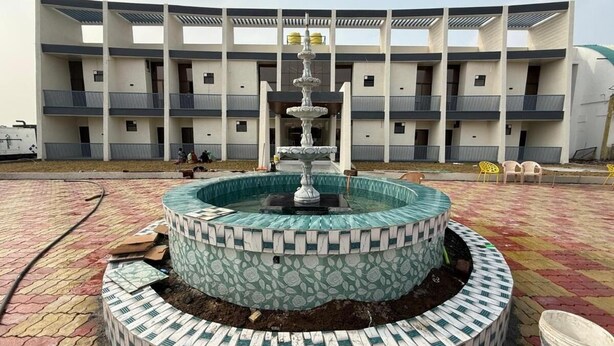 A multi-level fountain with a decorative top, surrounded by a patterned tiled base, in front of a modern building with balconies. - Hotel Madhuban (Khuldabad)