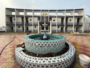 Une fontaine à plusieurs niveaux, surmontée d’un élément décoratif et entourée d’une base carrelée à motifs, devant un bâtiment moderne doté de balcons.