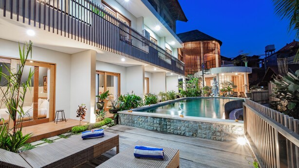 A modern outdoor pool area with a stone-lined edge, surrounded by lush greenery and a wooden deck. - Juli House (Ubud)