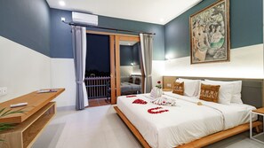 A hotel room with a bed, a desk, a balcony, and a painting on the wall. - Juli House (Ubud)