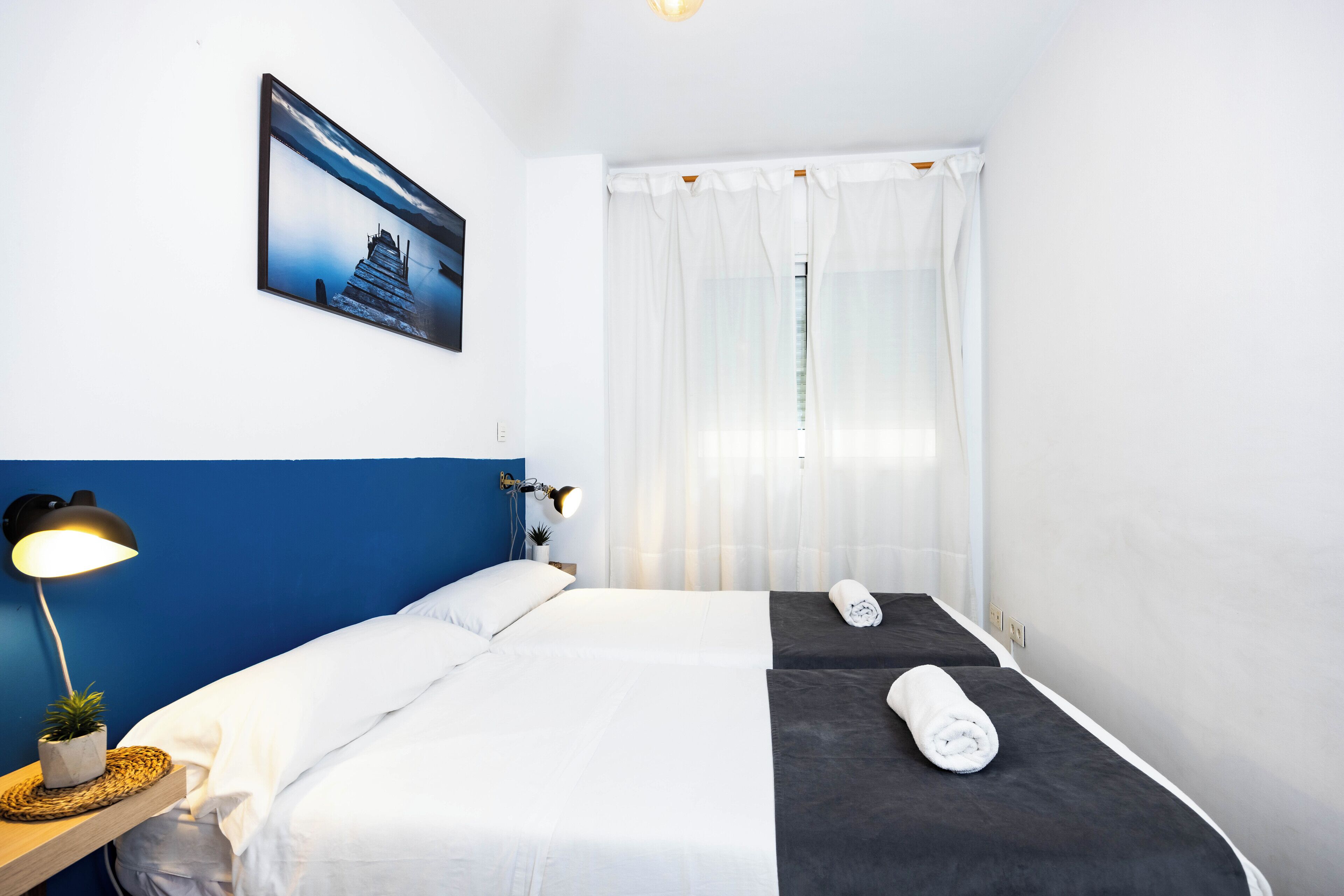 Habitació d'hotel amb llit, una làmpada de nit, una taula petita amb una planta i una finestra amb cortines.