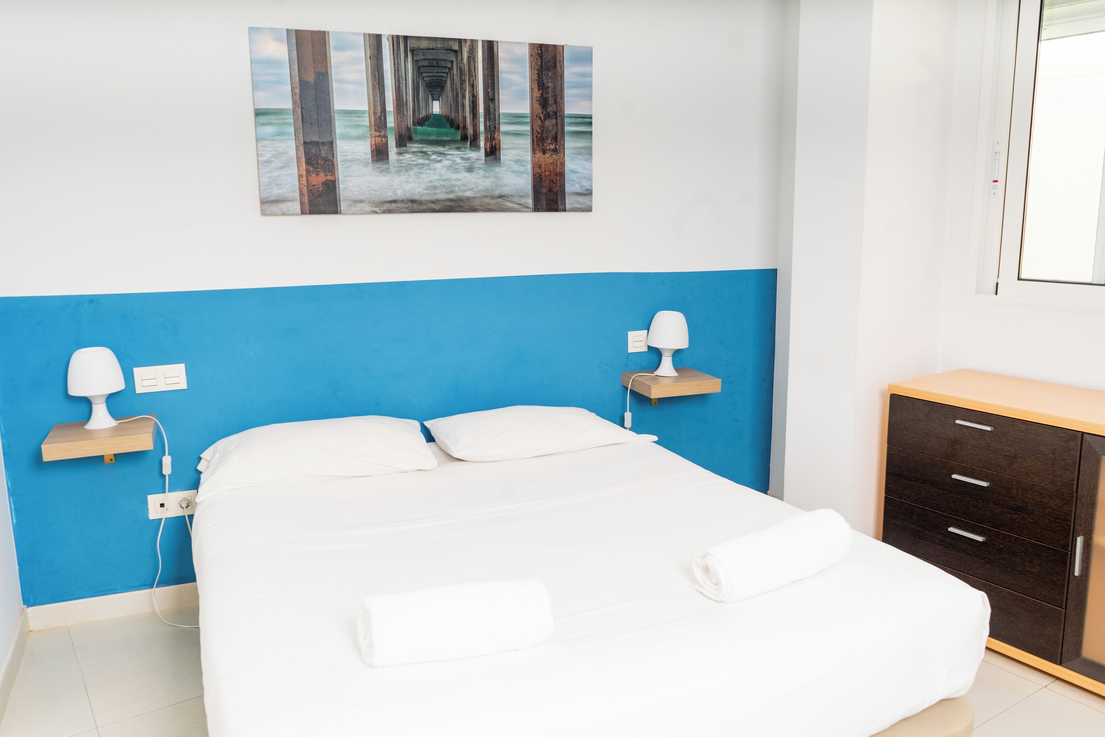 A bedroom with a bed, bedside lamps, a wooden nightstand, a wooden dresser, and a painting of a pier.