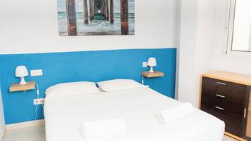 A bedroom with a bed, bedside lamps, a wooden nightstand, a wooden dresser, and a painting of a pier.
